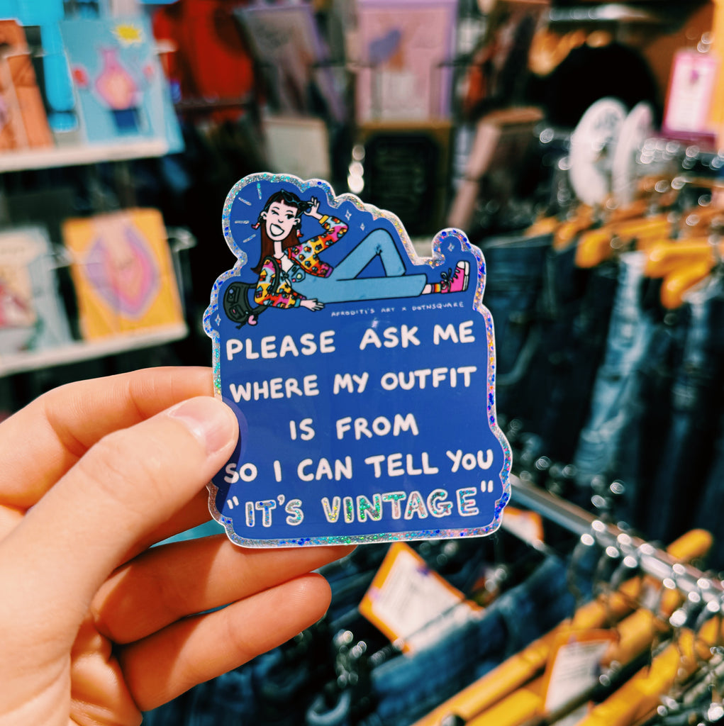 Hand holding a blue sticker with text "please ask me where my outfit is from so I can tell you it's vintage" in a clothing store