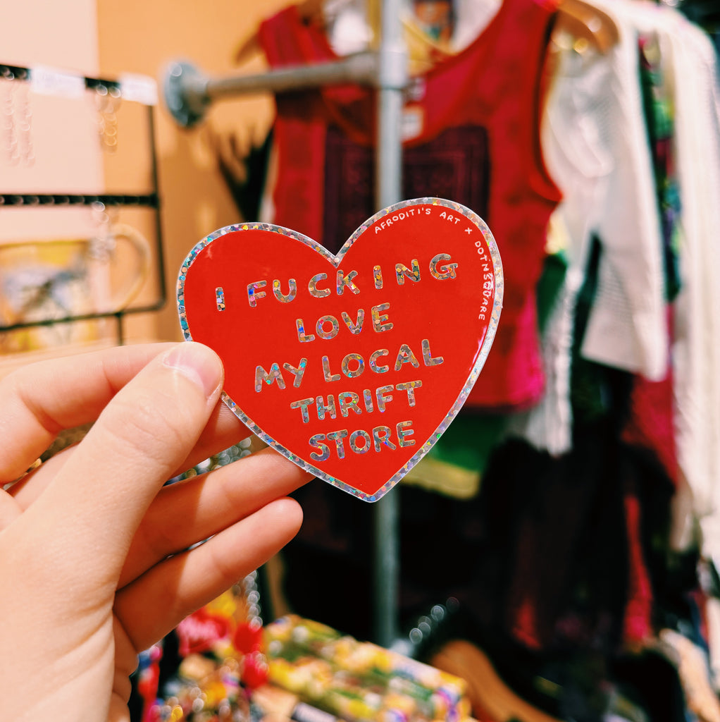 Heart-shaped red sticker with text "I fucking love my local thrift store" held in front of a clothing store background