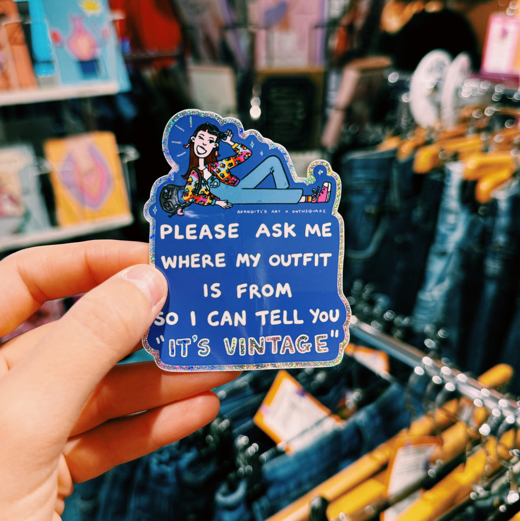 Hand holding a blue sticker with text "please ask me where my outfit is from so I can tell you it's vintage" in a store
