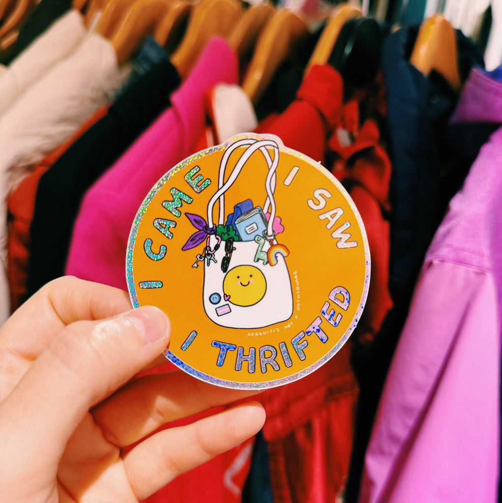 Hand holding a round sticker with text reading "I came I saw I thrifted" in front of a clothing rack.