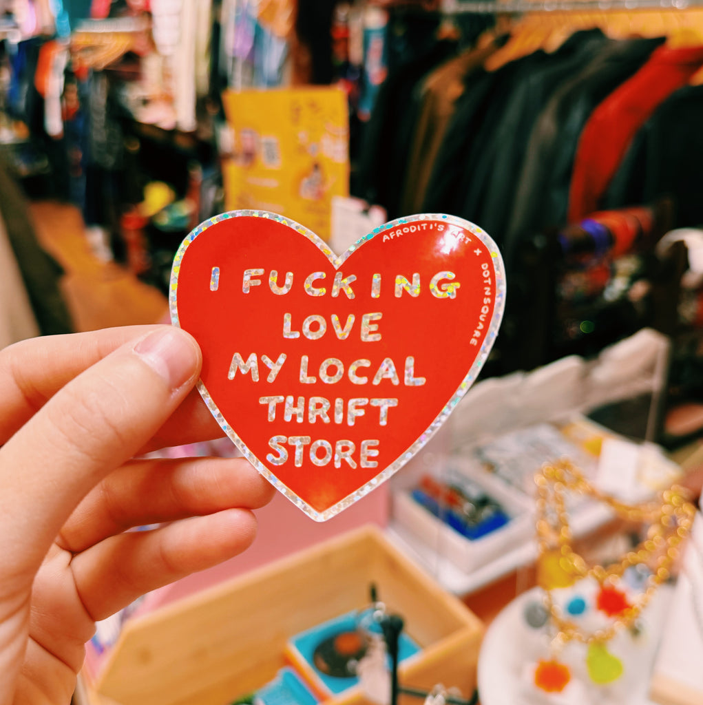 Heart-shaped sticker with text "I fucking love my local thrift store" held in front of a store interior.