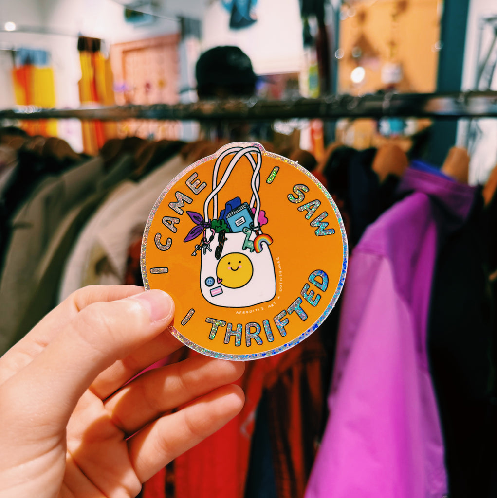 Hand holding a round orange sticker with text  reading "I came I saw I thrifted"