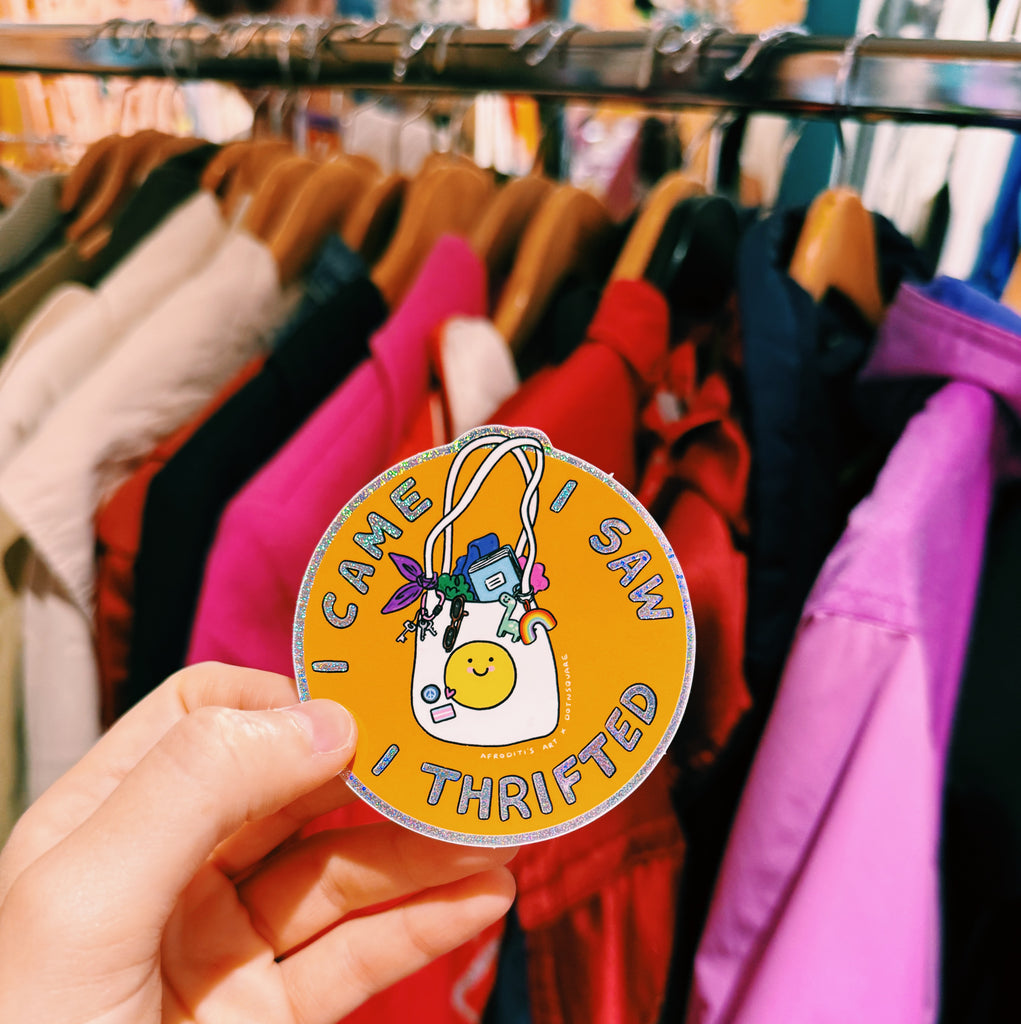 Hand holding a sticker with text reading "I came I saw I thrifted" in front of a clothing rack.