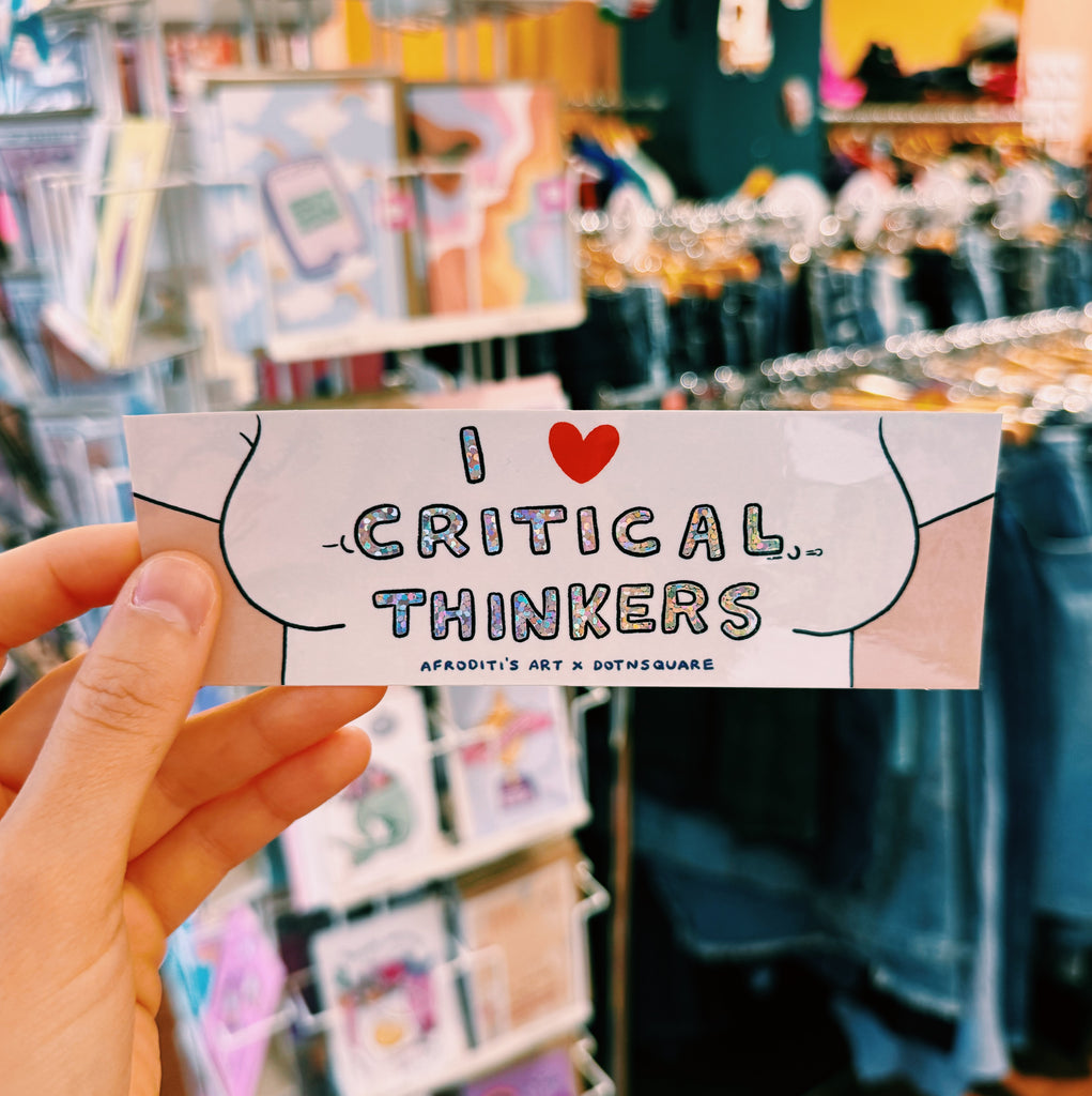 Sticker with 'I ❤️ Critical Thinkers' text held in front of a store 