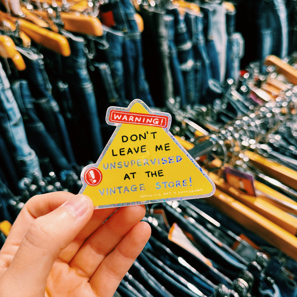 Hand holding a yellow warning sign sticker with text "Warning! Don't leave me unsupervised at the vintage store!" in front of a clothing rack.