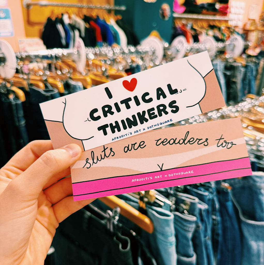 bookmark with text 'I ❤️ Critical Thinkers, sluts are readers too' held in front of a clothing store.
