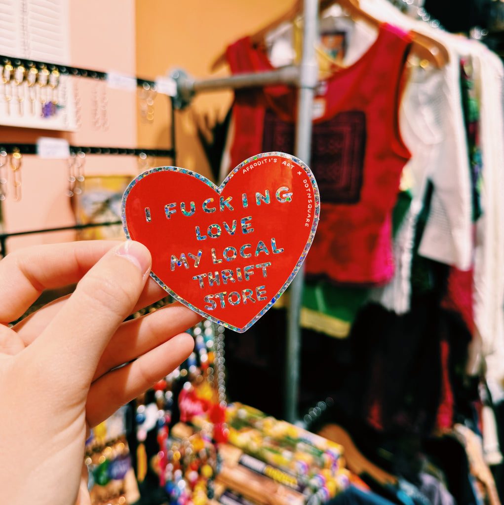 Red heart-shaped sign with text "I fucking love my local thrift store" held in front of a clothing store display.