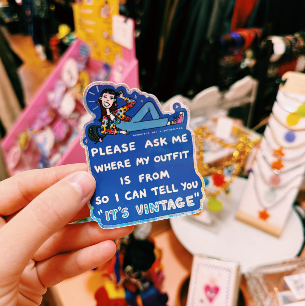 Hand holding a blue sticker with text "Please ask me where my outfit is from so I can tell you it's vintage" and a cartoon character, with a blurred background of a store.
