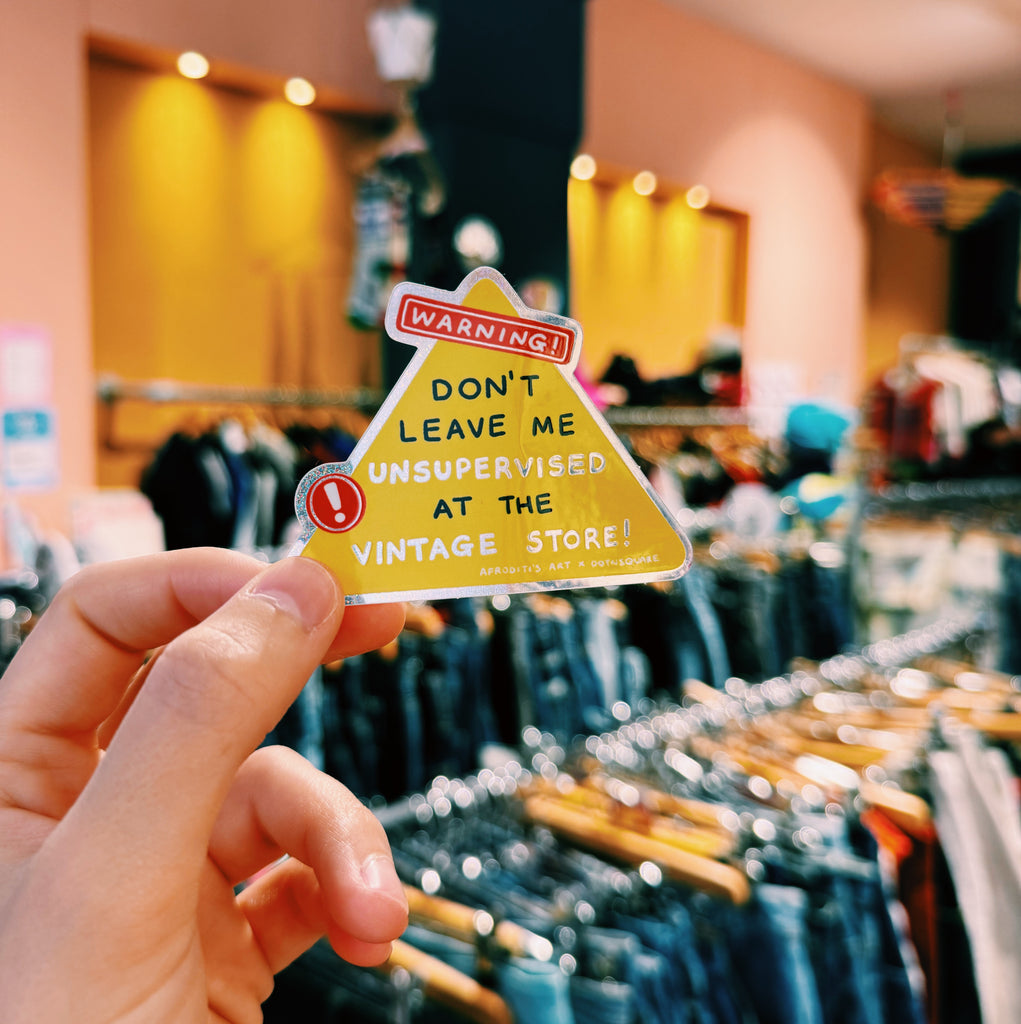 Hand holding a warning sign sticker reading "warning! Don't leave me unsupervised at the vintage store!" in front of a vintage store with clothing racks.