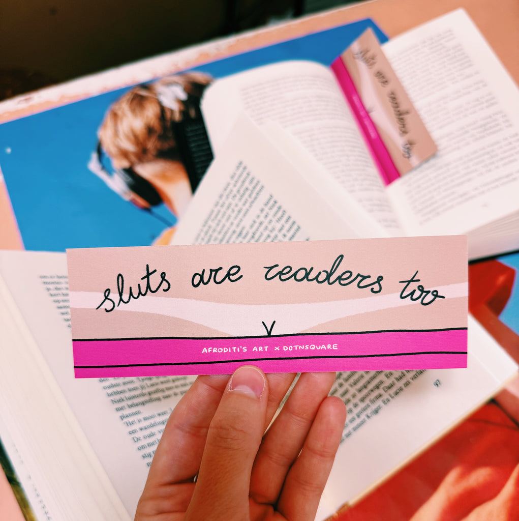 Hand holding a bookmark with text "sluts are readers too" over an open book