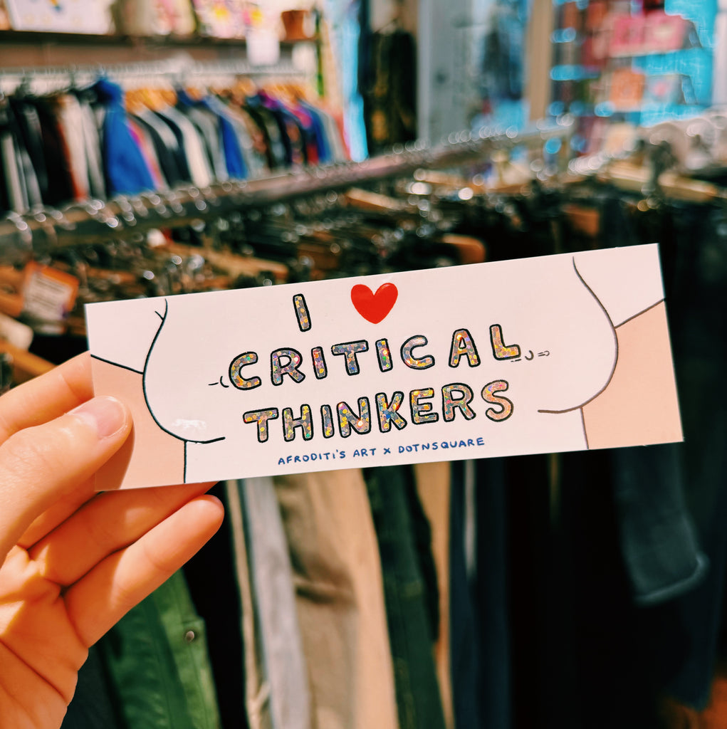 Hand holding a sticker with 'I ❤️ Critical Thinkers' text in a store