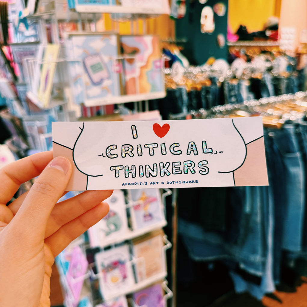 Hand holding a sticker with 'I ❤️ Critical Thinkers' text in glitter effect in a store